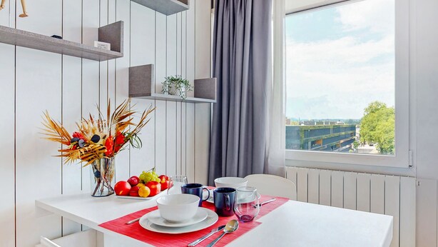 A dining area with a table set for two, a vase with dried flowers, a fruit plate, and a view of the city through a window. - Evi's Home Hotel & Apartments (Bern)