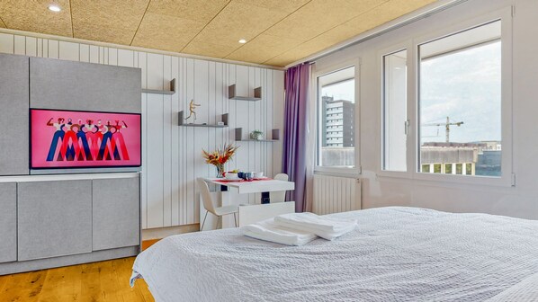 A modern hotel room with a large bed, a desk with a chair, a TV mounted on the wall, and a window with a view of the city. - Evi's Home Hotel & Apartments (Bern)