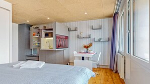 A modern bedroom with a bed, a desk, and a chair. There is a window with curtains, a microwave, and a shelf with decorative items. - Evi's Home Hotel & Apartments (Bern)