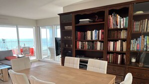 A spacious living area with a large wooden dining table surrounded by white chairs, a bookshelf filled with books, and a balcony with a view of the sea.