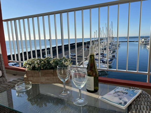 A balcony with a glass table, a bottle of wine, two wine glasses, and a magazine. - Beautiful 2-BR, A/C, Parking, Near Beach and Shops (Cap d'Agde)