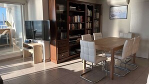 A modern living room with a wooden dining table, white cushioned chairs, a large bookshelf, a flat-screen TV, and a large window with a view of the sea.