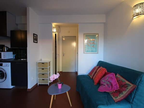 A living room with a teal sofa, a small round table with a pink flower, a washing machine, and a microwave.