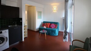 Un espacio habitable compacto con kitchenette, un sofá con cojines de colores, una pequeña mesa redonda y un aplique de pared.