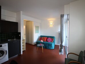 A compact living space with a kitchenette, a sofa with colorful cushions, a small round table, and a wall-mounted light fixture.
