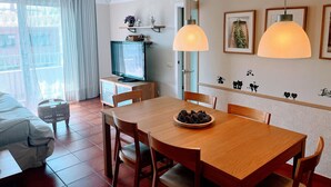 A dining area with a wooden table, chairs, a television on a low stand, and a decorative wall with framed pictures.