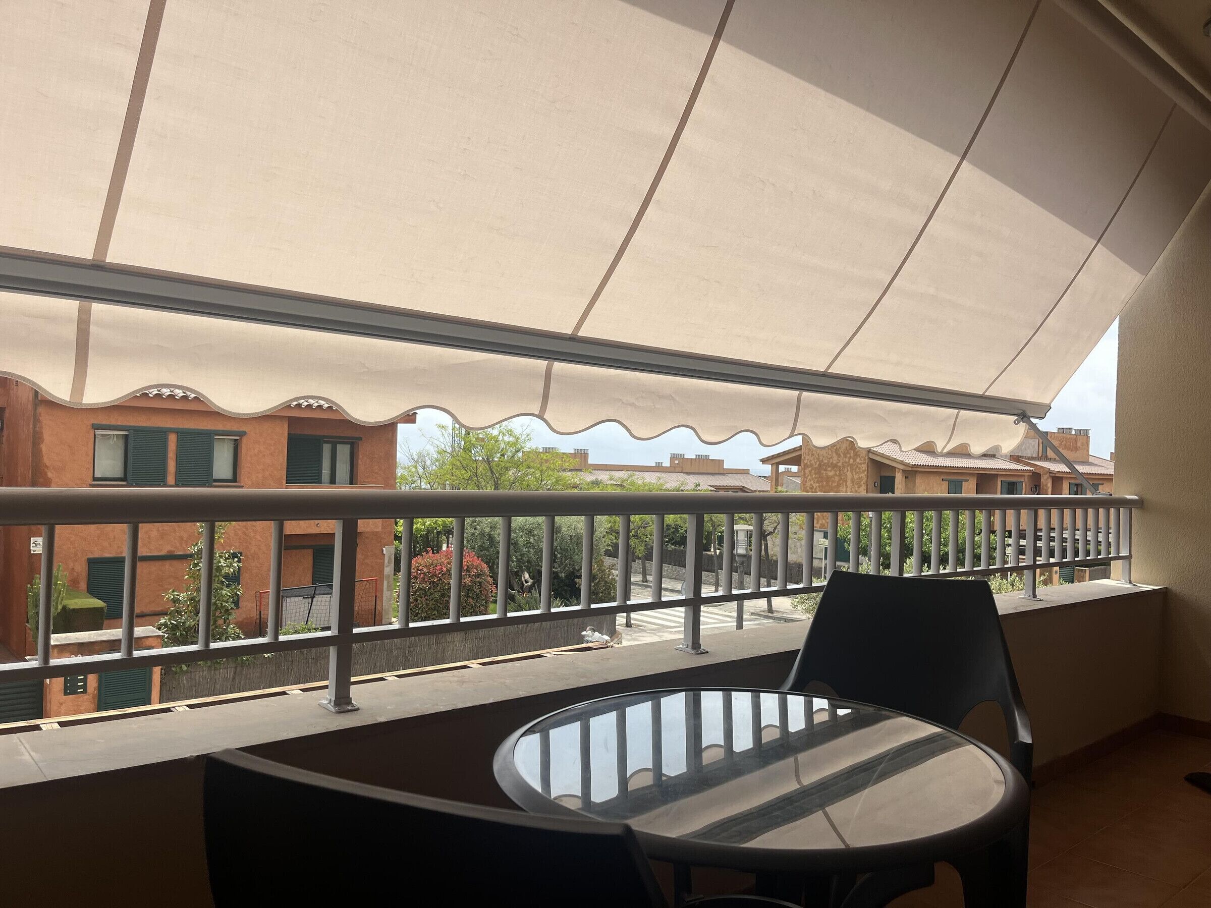 A balcony with a table and chairs, a view of neighboring buildings, and a large awning providing shade.