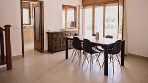 A dining area with a wooden table and chairs, a kitchen area with cabinets, and large windows with curtains. - Coastal Villa with Private Pool, Steps from Beach, 4 Bedrooms, BBQ, Wi-Fi, Family-Friendly (L'Ametlla de Mar)