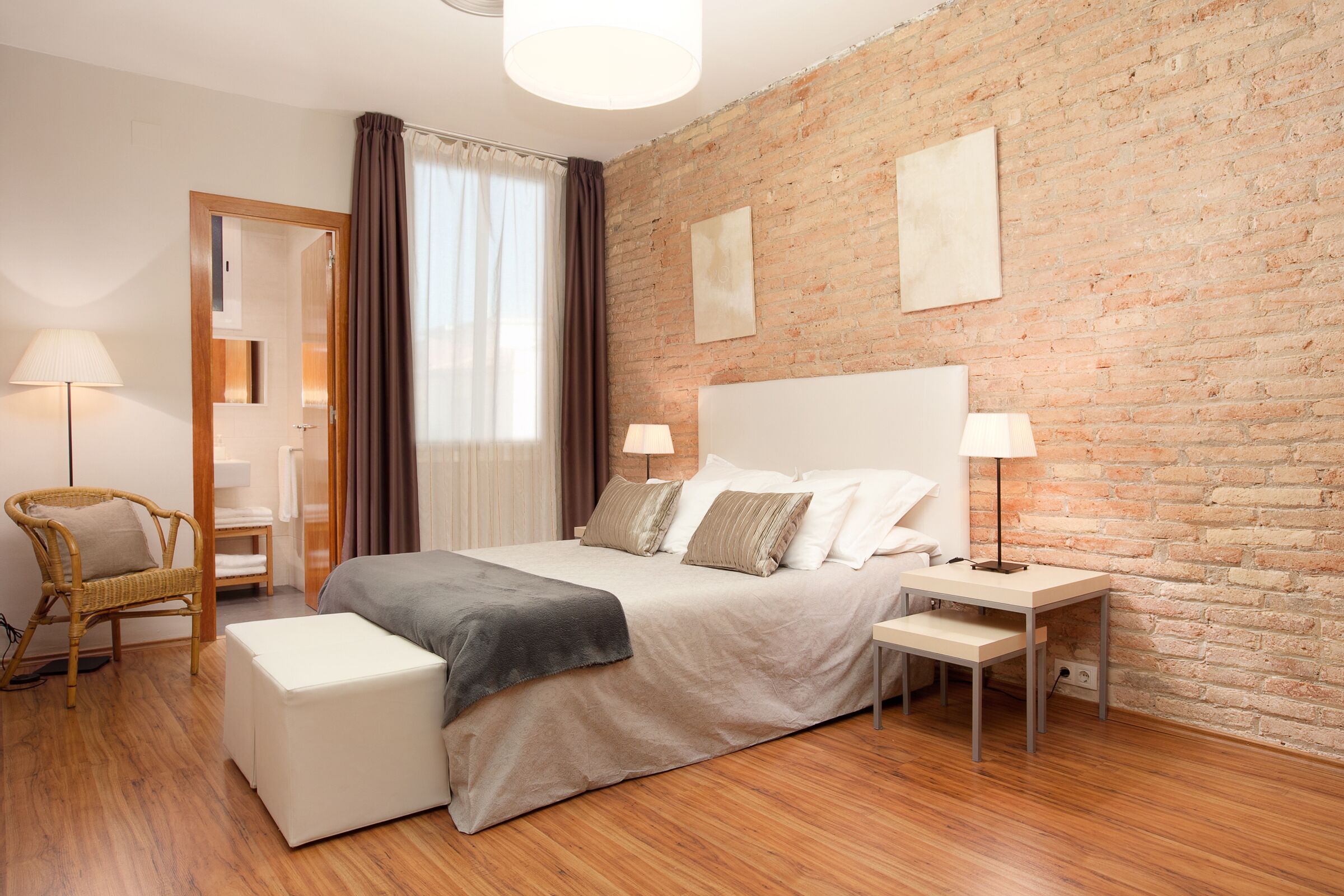 A modern hotel room with a brick wall, a large bed with a headboard, two bedside tables with lamps, a wooden floor, a wicker chair, and a small table.