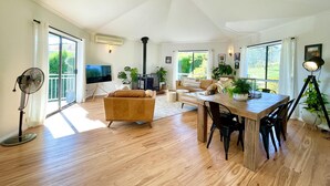 Ein großzügiges Wohnzimmer mit Holzboden, einer Couch, einem Esstisch mit Stühlen, einem Fernseher und einem großen Fenster mit Blick auf Grünflächen.