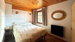 A bedroom with a wooden ceiling, a bed with a floral bedspread, a bedside table with a lamp, a window with curtains, and a wall-mounted TV. - 4-sided Cottage 80m² near lake with garage (Matemale)