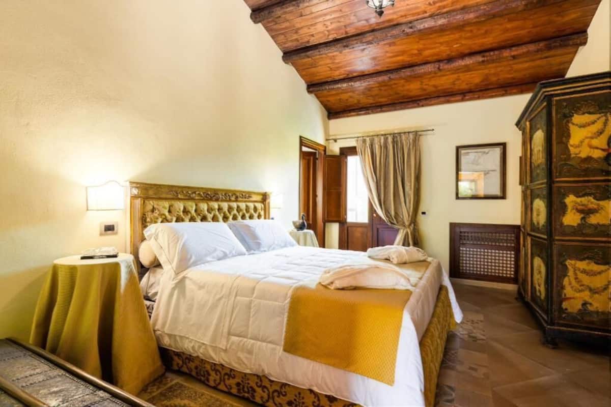 A bedroom with a large bed, a wooden ceiling, a nightstand, a dresser, and a window with curtains.