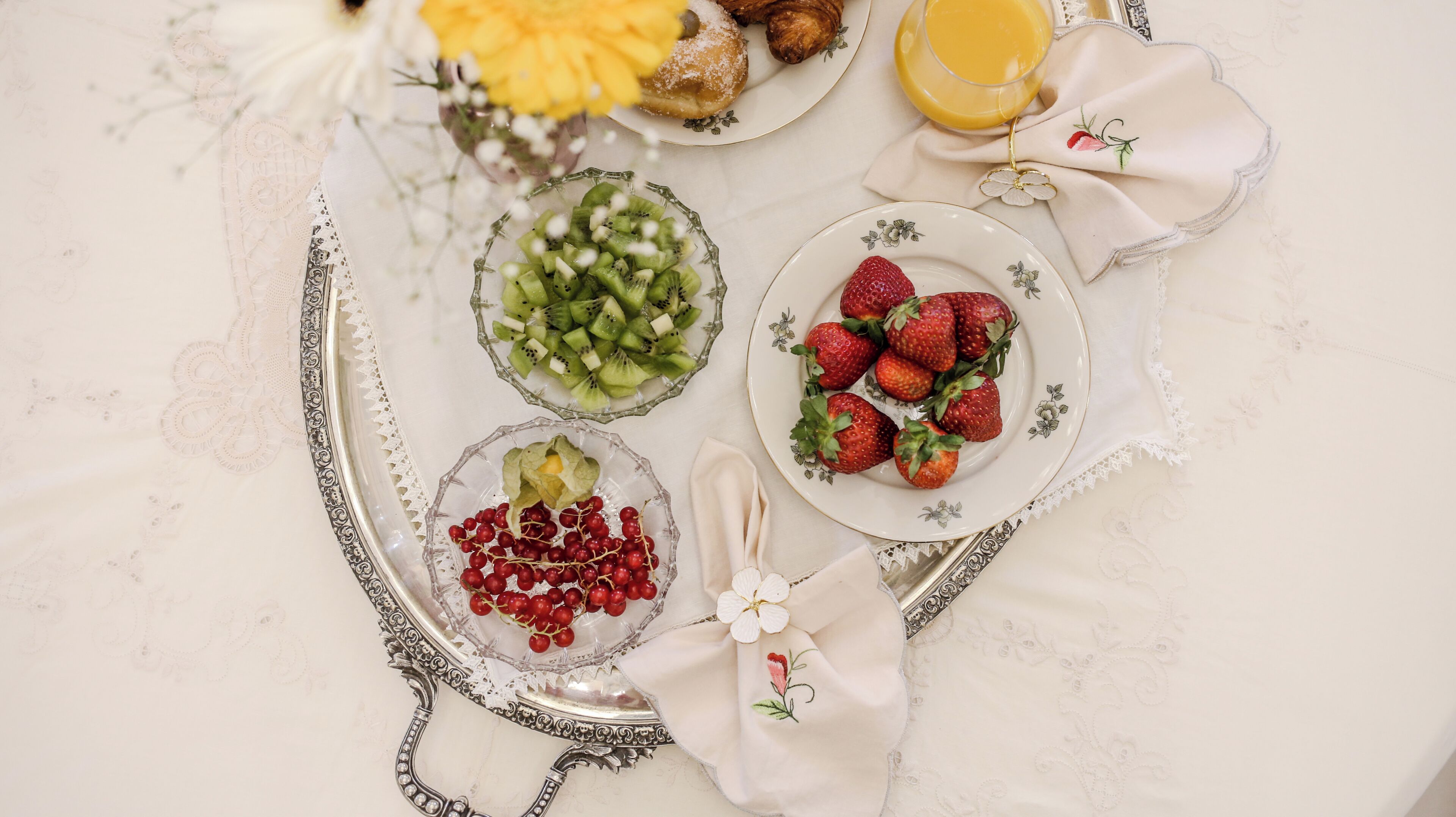A table with a variety of fruits and a glass of orange juice, arranged on a white tablecloth with lace details.