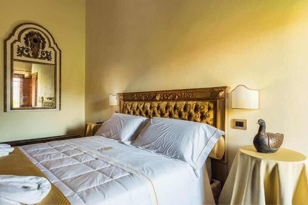 Ein Hotelzimmer mit einem großen Bett, einem Kopfteil mit aufwendigen Schnitzereien, einem runden Tisch mit einer verzierten Vogeldekoration und einem an der Wand befestigten Spiegel.