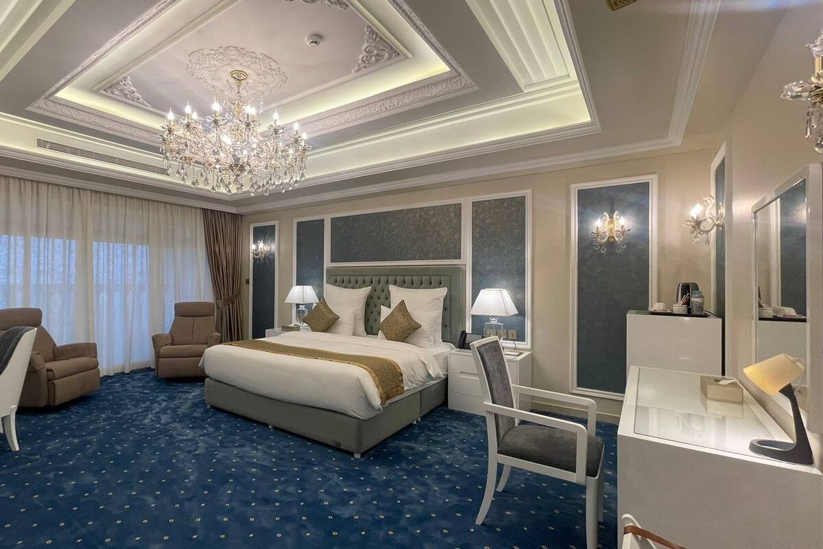A hotel room with a large bed, two armchairs, a desk, a chandelier, and a large mirror.