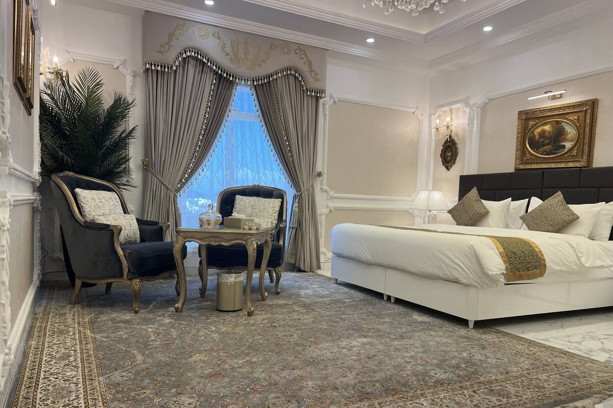 A luxurious bedroom with a large bed, two armchairs, a small table, and a large window with curtains.