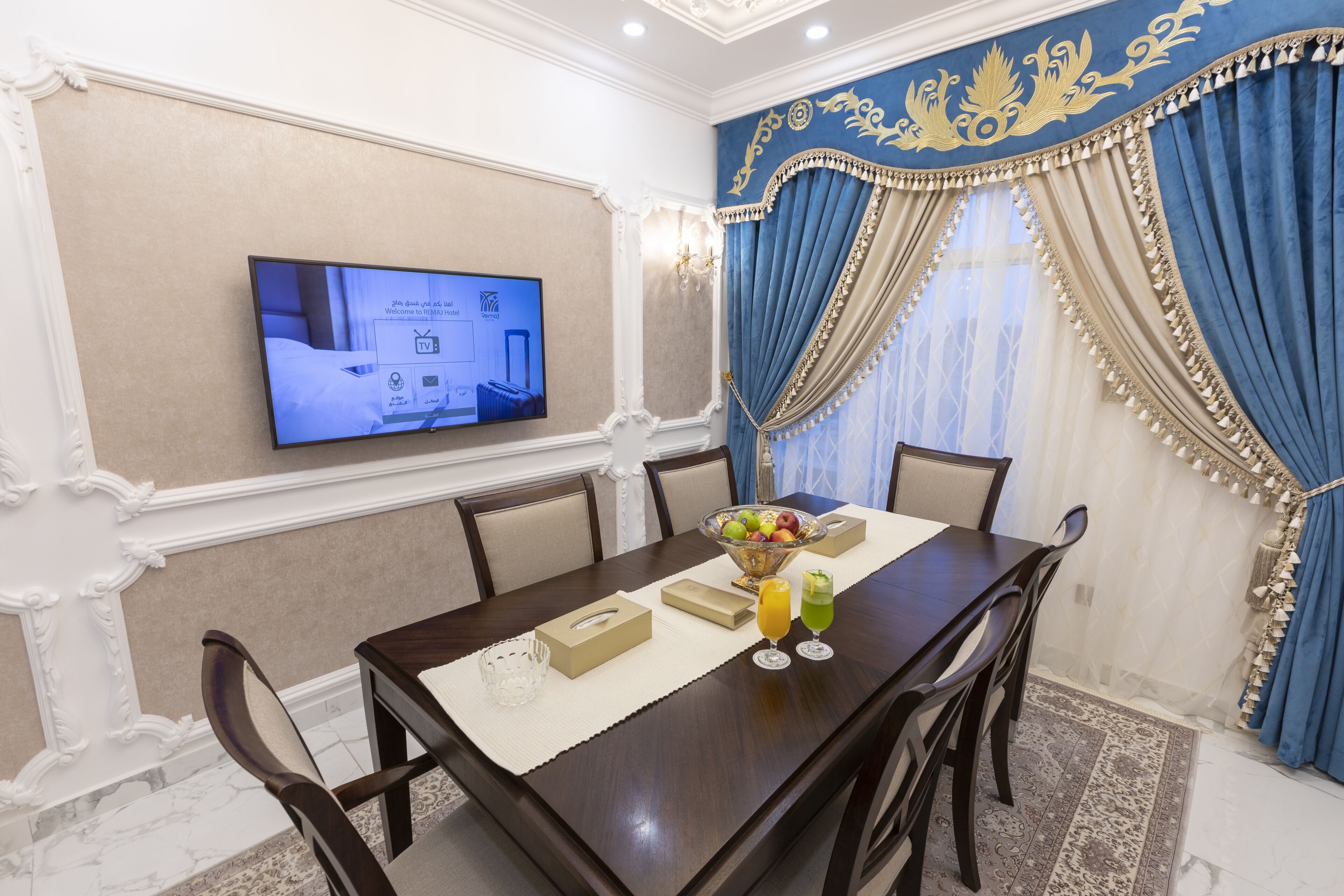 A hotel dining room with a wooden table, chairs, a TV screen, a fruit bowl, and a decorative wall.