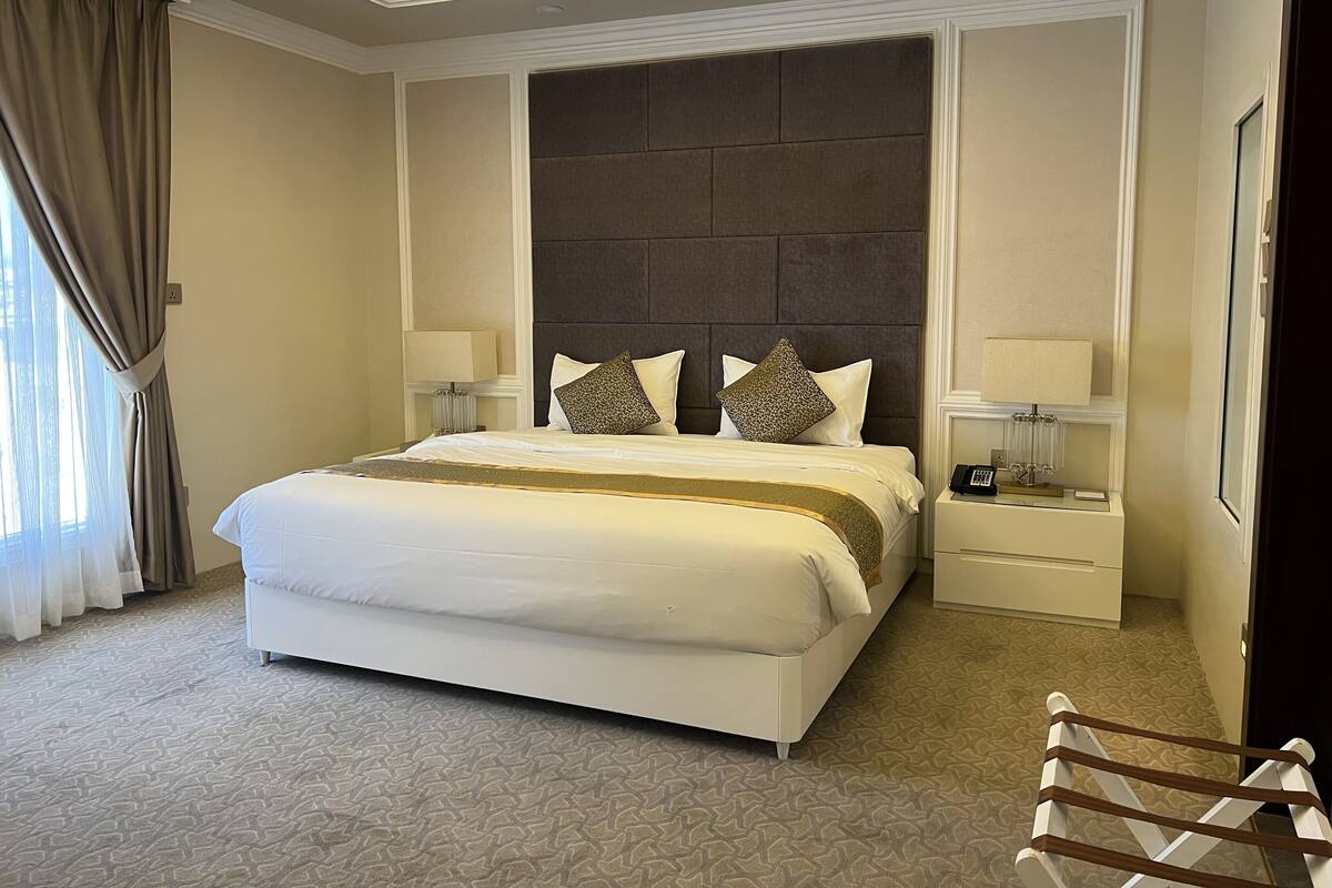 A hotel room with a large bed, two bedside tables, a headboard, and a view of the outdoors.