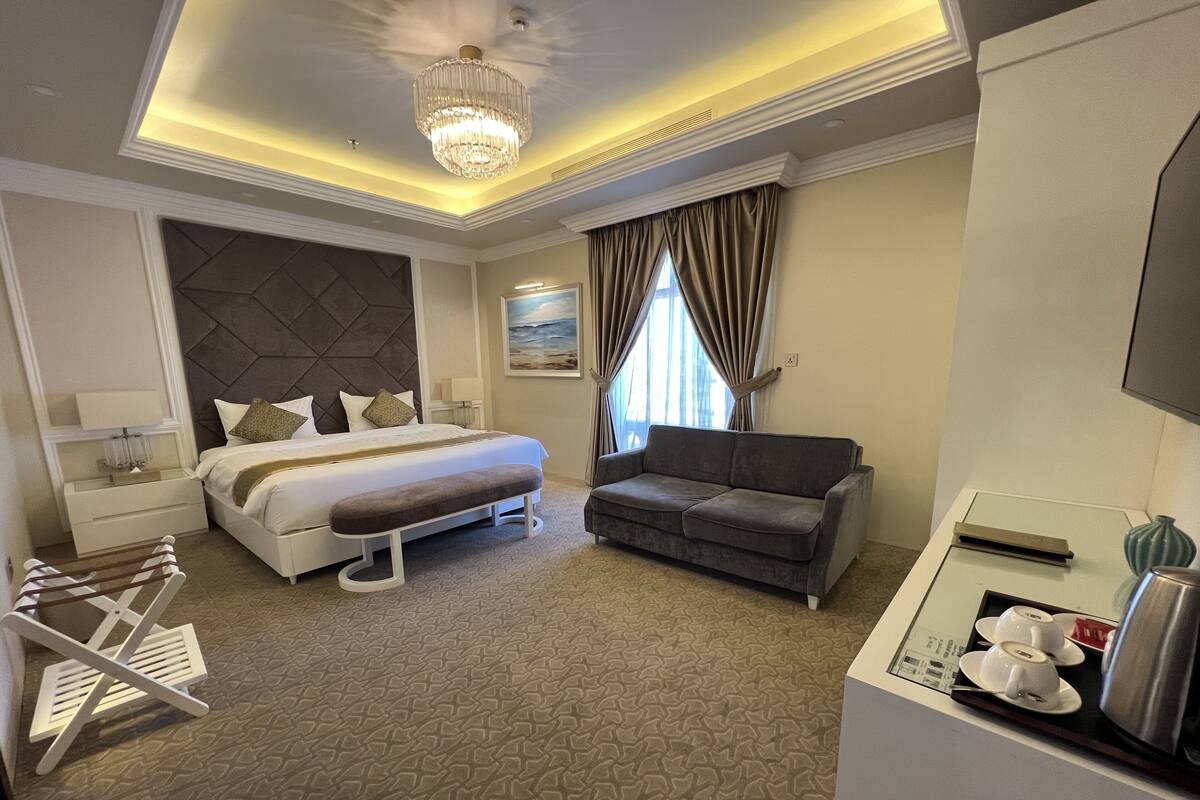 A hotel room with a bed, a sofa, a desk with a kettle, and a window with curtains.