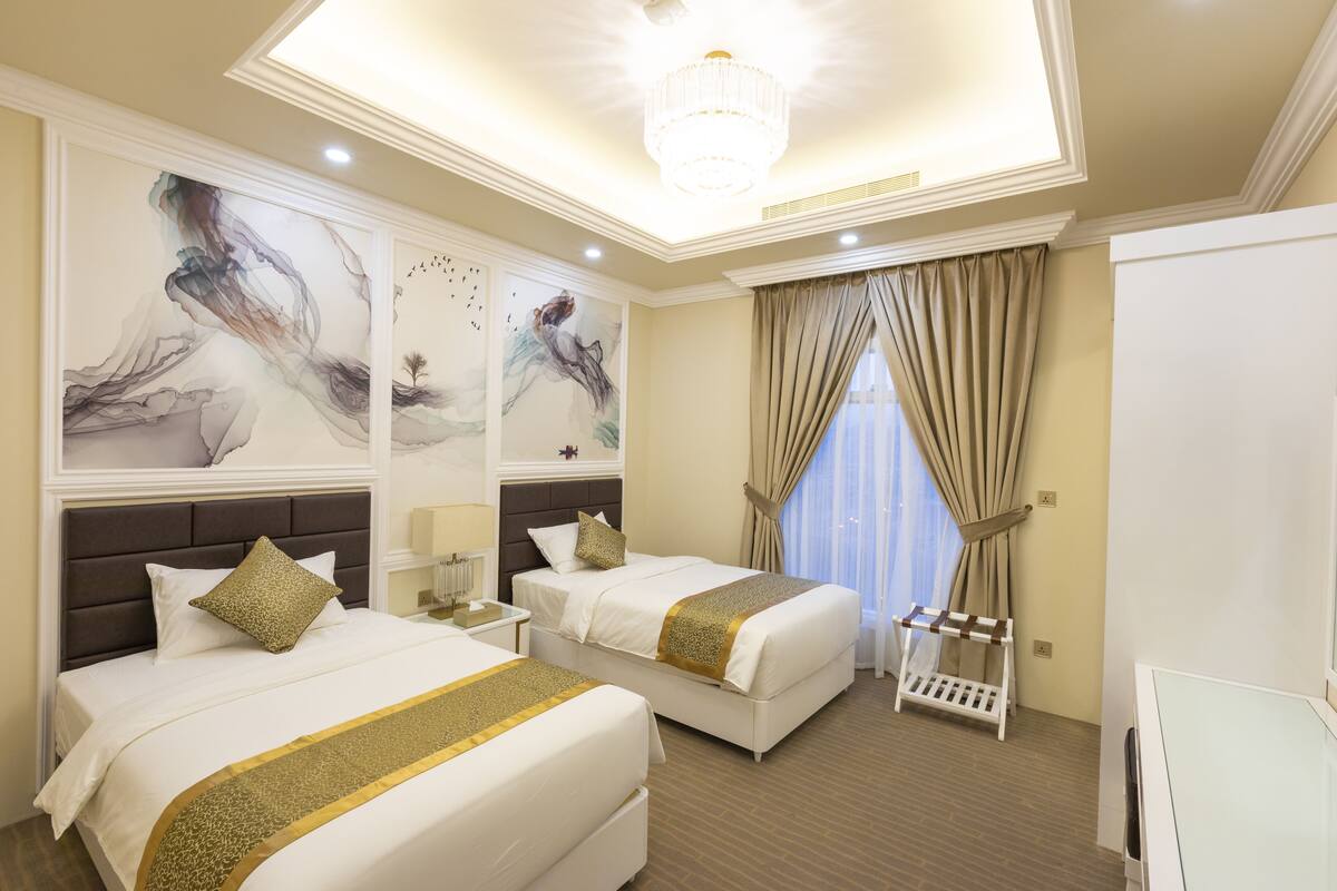 A hotel room with two beds, a chandelier, artwork on the walls, and a view of the outdoors through the window.