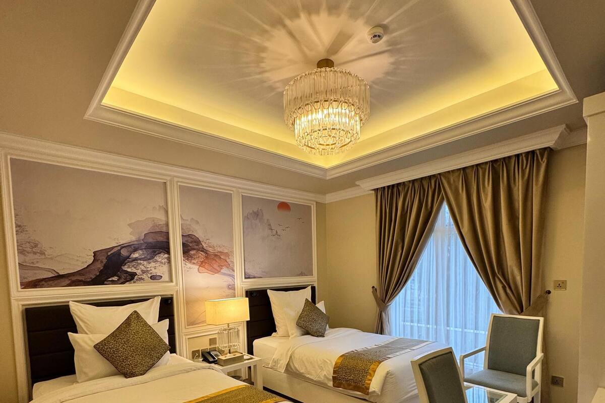 A hotel room with two beds, a chandelier, artwork on the walls, and a patterned carpet.