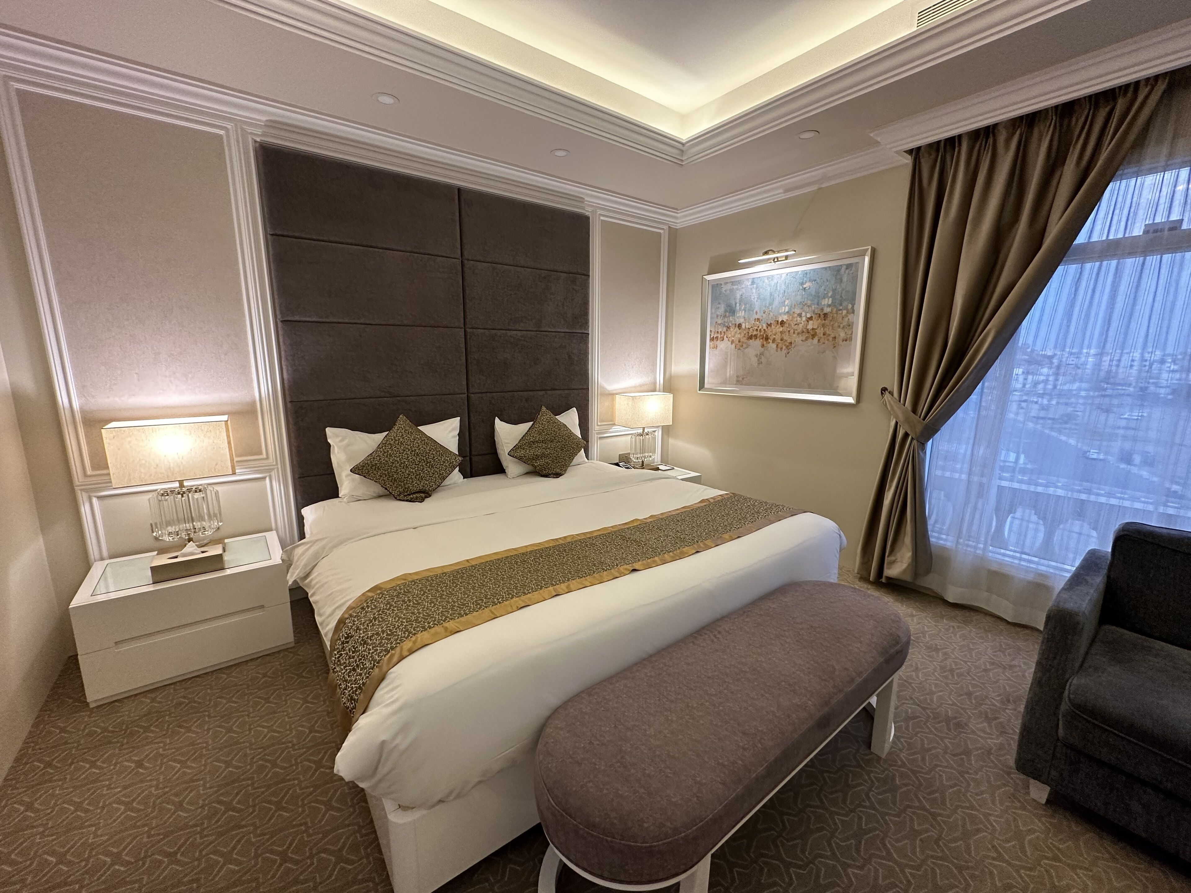 Habitación de hotel con una cama grande, dos mesitas de noche con lámparas, un banco, un sillón gris y un ventanal con cortinas.