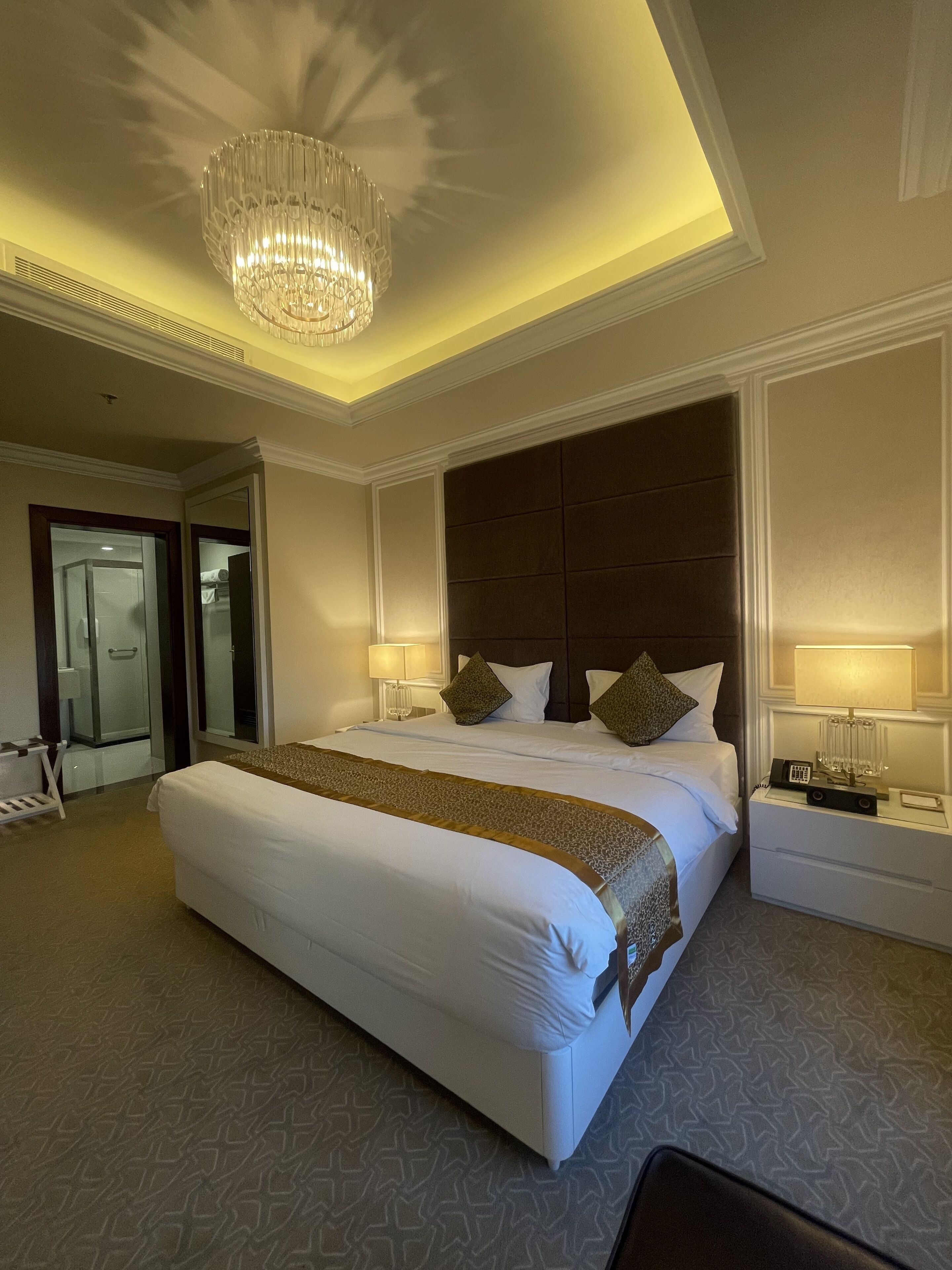 A spacious hotel room with a large bed, a chandelier, a nightstand, and a bathroom visible through an open door.