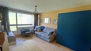 Huoneessa on puulattia, harmaa sohva koristetyynyineen, sininen vaatekaappi ja työpöytä tuolilla.