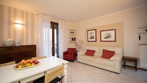 A living room with a dining table, a red armchair, a white sofa with red cushions, and a bookshelf. - Appartamento Maria Cristina (Polpenazze del Garda)