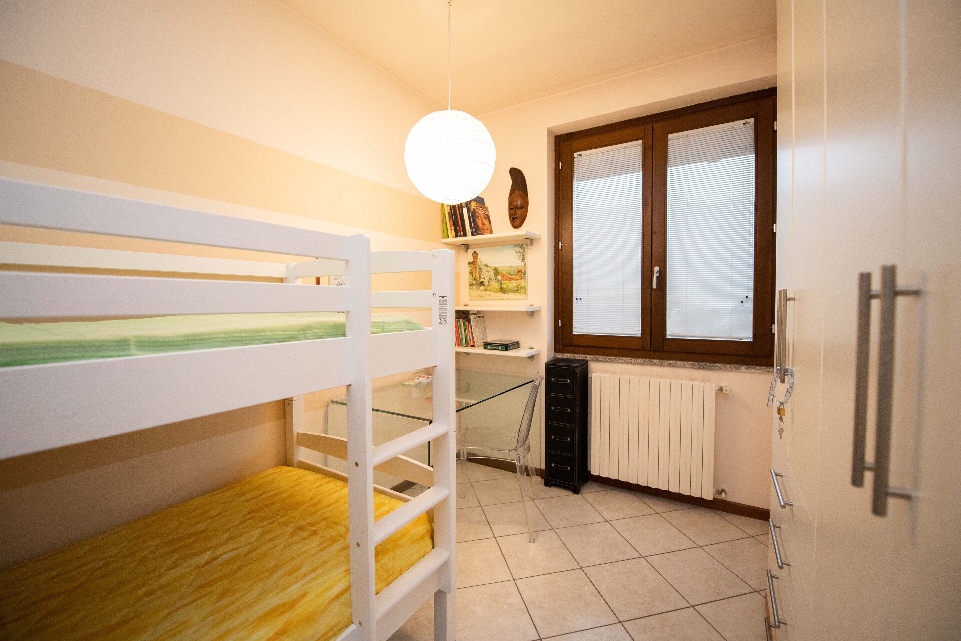 A bedroom with bunk beds, a desk, a chair, a radiator, and a window with blinds.