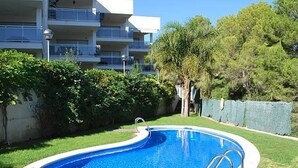 A swimming pool with a wooden deck, surrounded by a grassy area and a low wall. - Ground Floor Beach Apartment with Pool Access in Costa Dorada (L'Ametlla de Mar)