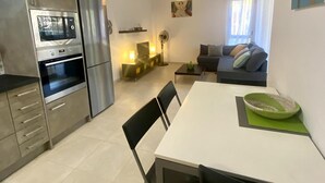 A modern kitchen with a built-in oven and microwave, a dining area with a white table and chairs, and a living area with a sofa and a TV. - Ground Floor Beach Apartment with Pool Access in Costa Dorada (L'Ametlla de Mar)