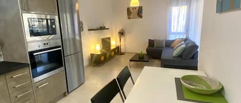 Une cuisine moderne dotée d’un four et d’un micro-ondes encastrés, un coin repas avec une table et des chaises blanches, et un salon avec un canapé et un téléviseur.
