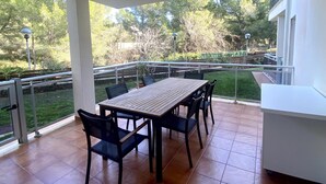 A covered patio with a wooden table and chairs, a small white cabinet, and a view of a garden with trees and a fence. - Ground Floor Beach Apartment with Pool Access in Costa Dorada (L'Ametlla de Mar)