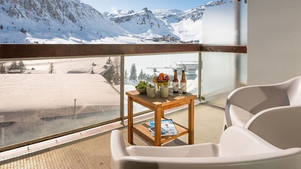 A balcony with a view of snow-covered mountains, a wooden table with bottles and fruits, and a white chair. - Premium Apartment in Tignes for 6 ppl with WIFI (Tignes)
