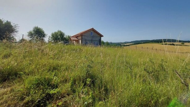 Una casa rural con techo rojo, rodeada de hierba alta y campos.