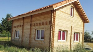 Una cabaña de madera con techo de tejas, dos ventanas con contraventanas y un paisaje natural circundante.
