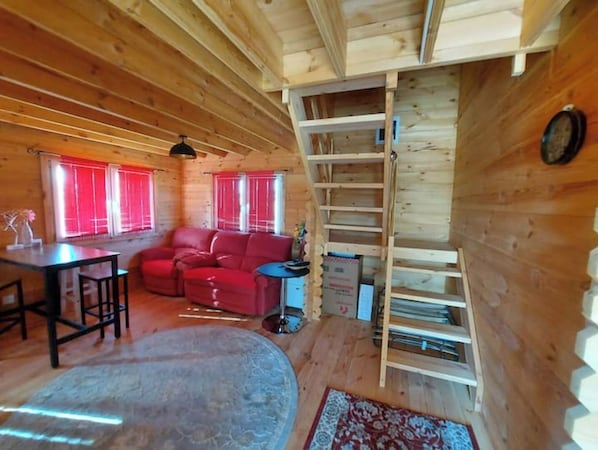 A wooden cabin interior with a sofa, dining table, and a ladder leading to a loft. - Pretty wooden house with large enclosed garden. (Lafage)