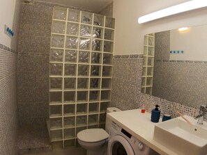 Kylpyhuone, jossa on laatoitettu suihkukaappi, wc, pesukone ja peilillä varustettu pesuallas.