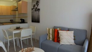 Een knus woongedeelte met een grijze bank, een klein rond tafeltje met decoratie, een witte eettafel met stoelen, en een keukenhoek met kasten en een koffieapparaat.