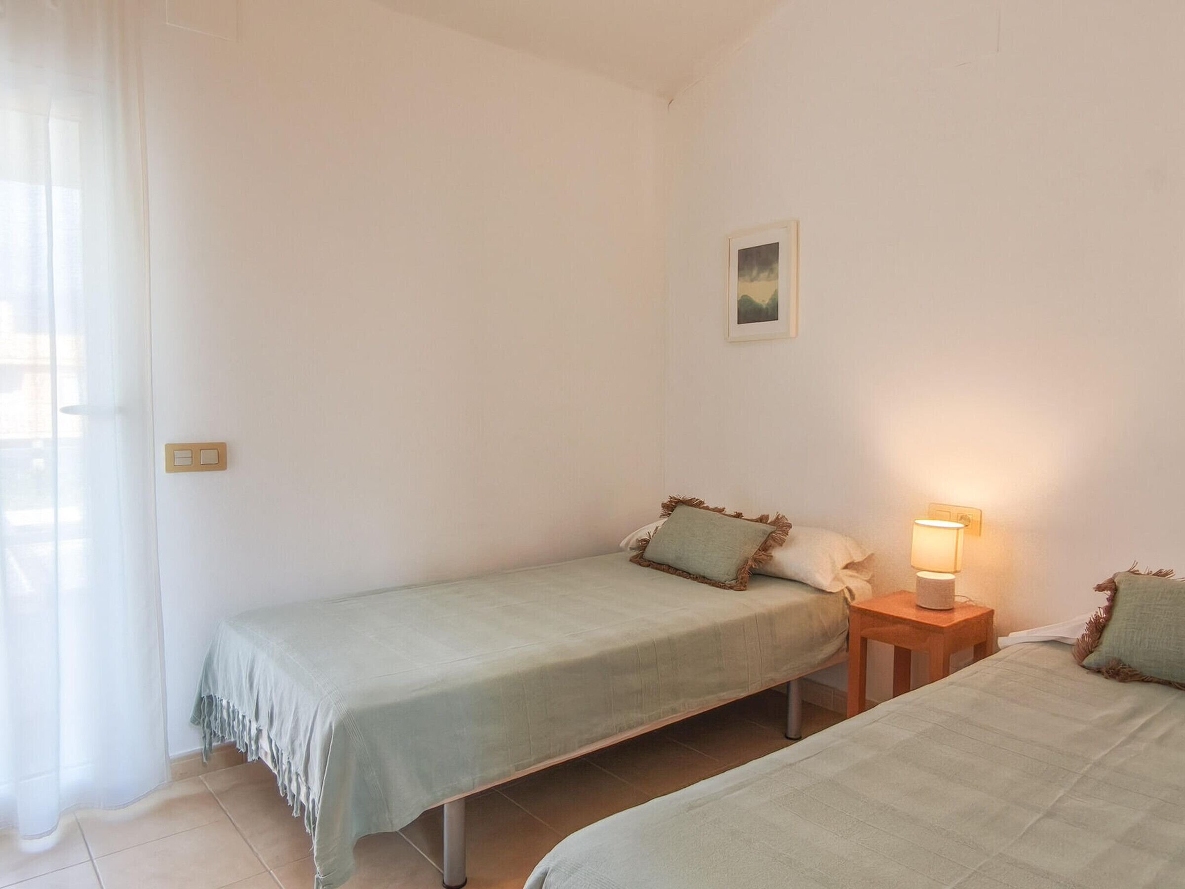 A small, clean room with two single beds, a bedside table with a lamp, and a window with sheer curtains.