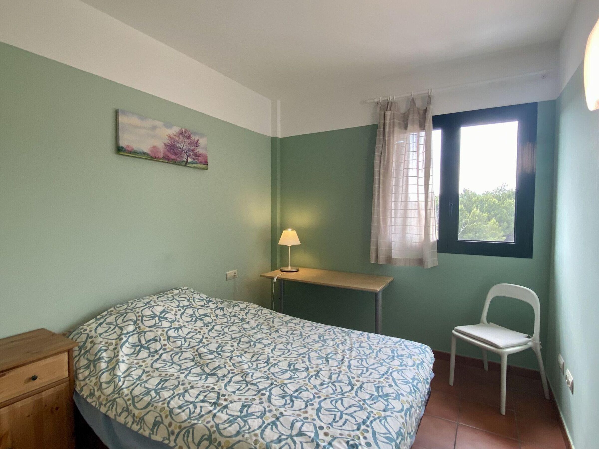 Un dormitorio con cama, escritorio, silla, lámpara y ventana con cortinas.