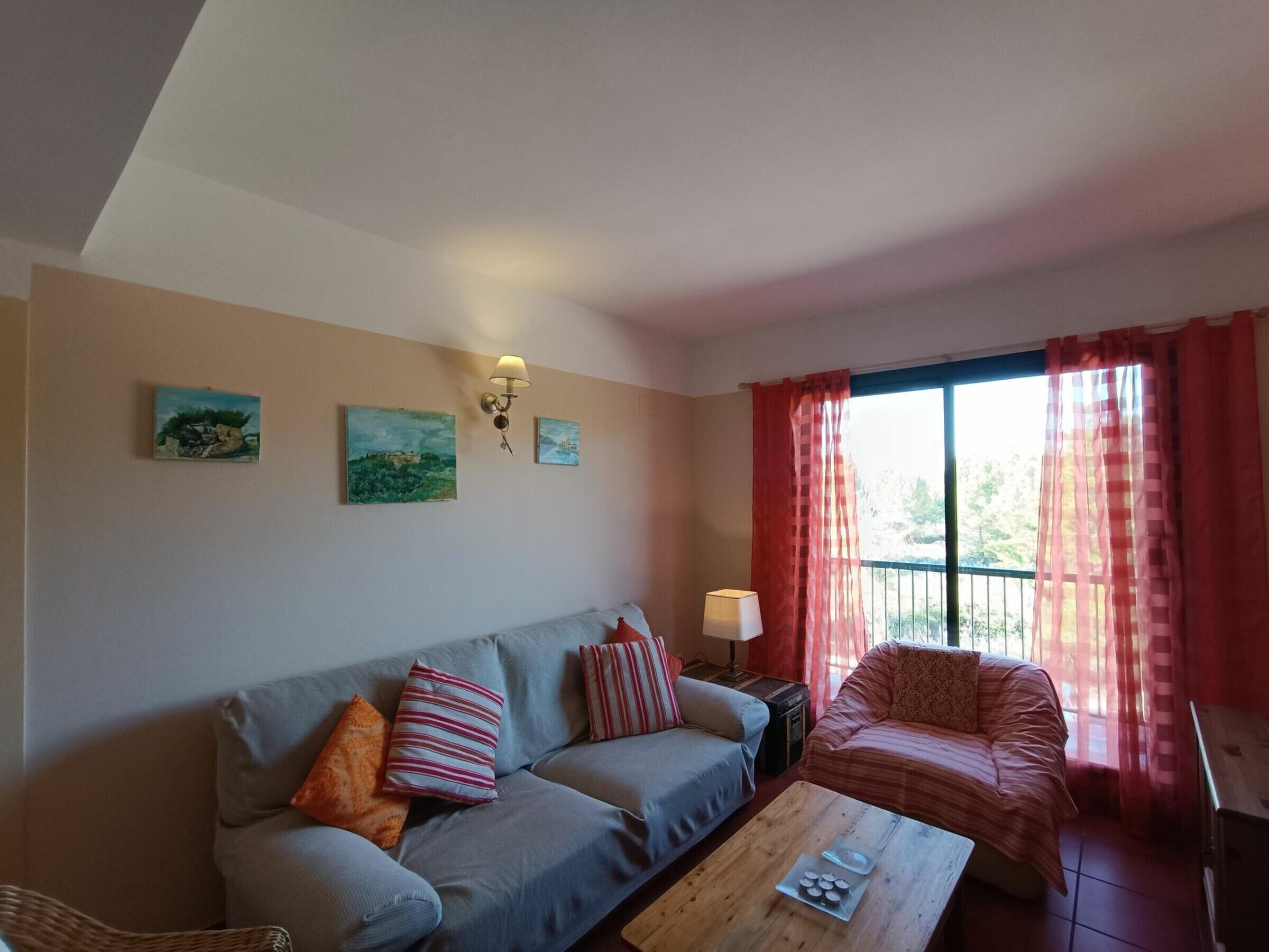 A living room with a sofa, a coffee table, and a window with curtains. There are wall-mounted lamps and paintings on the wall.