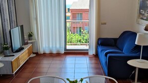 Ein Wohnzimmer mit Fernseher, einer Couch, einem Esstisch und Blick auf ein Gebäude durch ein Fenster.