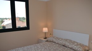 A bedroom with a bed, a nightstand, a lamp, and a window with a view of buildings and trees.