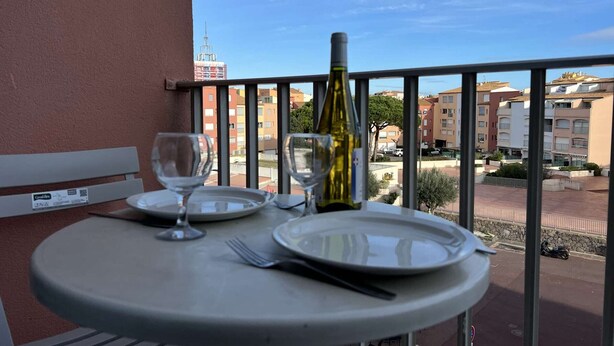 A balcony table with a bottle of wine, two wine glasses, and a plate, overlooking a residential area with buildings and a clear sky. - Charming Studio with Pool, Balcony, and Central Location for 2 in Agde (Cap d'Agde)