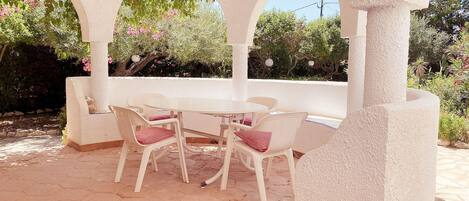 An outdoor patio with a table and chairs, surrounded by white arches and a garden with various plants and trees.