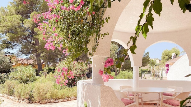 A covered outdoor seating area with a table and chairs, surrounded by lush greenery and flowering plants. - Charming Rustic Villa with Pool & Mediterranean Garden (L'Ametlla de Mar)