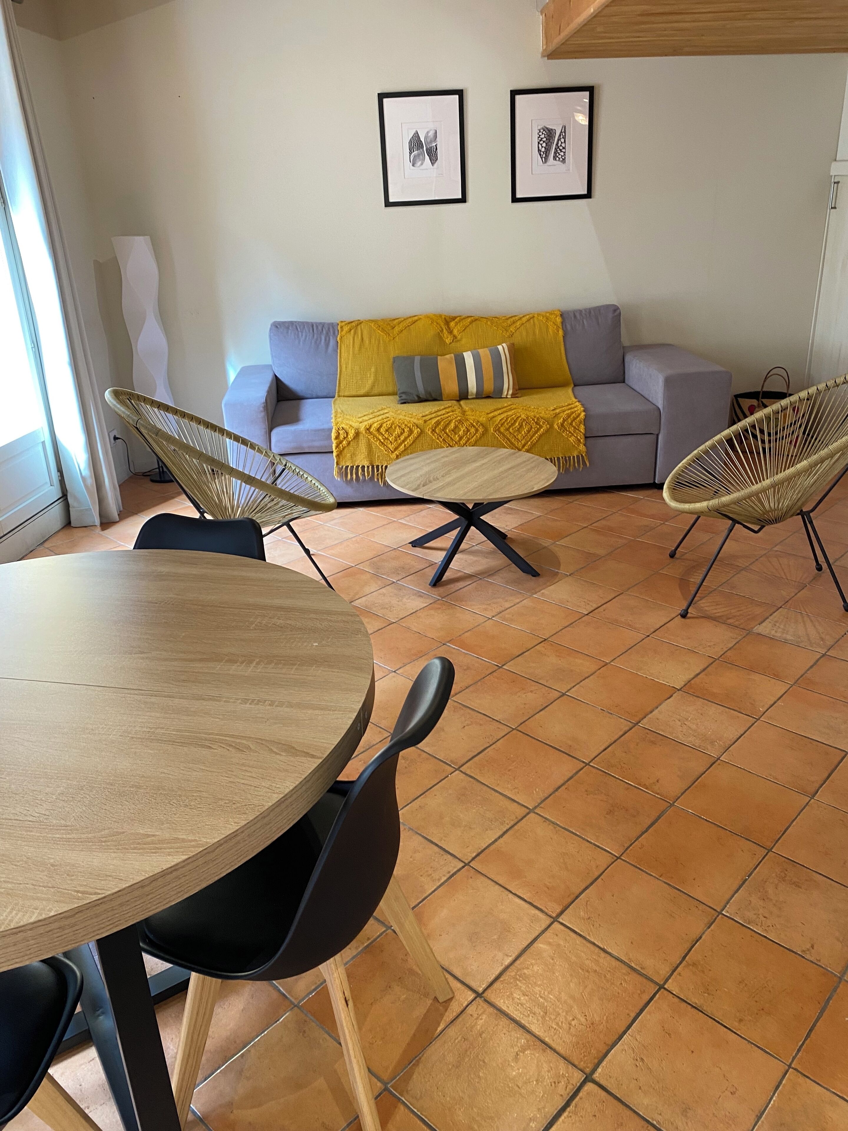 Un salon moderne avec une table ronde en bois, deux chaises noires, un canapé gris avec un plaid jaune, et deux œuvres d’art abstraites encadrées accrochées au mur.