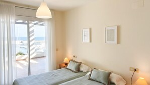 Un dormitorio con una cama, dos lámparas de noche, vista al mar a través de una puerta corredera y dos cuadros en la pared.